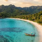 Aerial view of Koh Pha Ngan, Thailand, showing turquoise water, white-sand beaches, coconut palms, and a traditional long-tail fishing boat.
