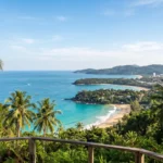 Panoramic view of Phuket's coastline from Karon Viewpoint, featuring turquoise Andaman Sea waters, tropical beaches, lush green hills, and coconut palm trees.